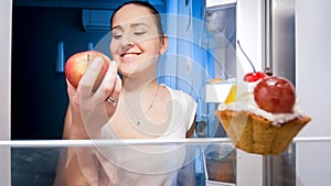 Portrait of smiling young woman taking apple out of refrgerator for late night snack