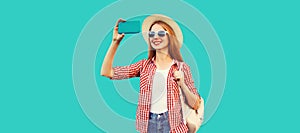 Portrait of smiling young woman taking selfie with smartphone wearing summer straw hat and backpack  on blue background