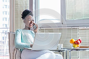 Portrait of a smiling young woman talking on mobile phone and working on laptop at home. Attractive young African woman working on