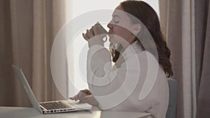 Portrait of smiling young woman using laptop and drinking coffee. Beautiful brunette Caucasian woman enjoying silent