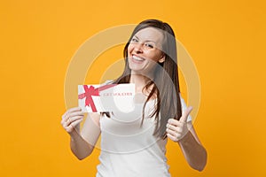 Portrait of smiling young woman in white casual clothes showing thumb up, holding gift certificate isolated on bright