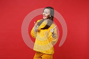 Portrait of smiling young woman in yellow fur sweater pointing index fingers on camera isolated on bright red wall