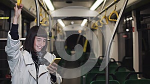 Portrait of smilling woman in headphones holds the handrail, listening to music and browsing on mobile phone in public