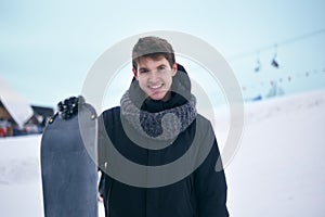 Portrait of snowboarder. Handsome man in ski suit is holding a snowboard, looking at camera and smiling. Man in the mountains