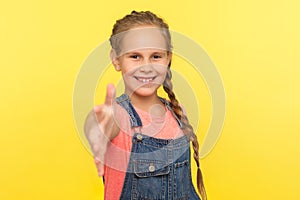 Portrait of sociable happy little girl with braid in denim overalls smiling friendly and giving hand to handshake