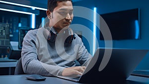 Portrait of Software Developer / Hacker / Programmer Sitting at His Desk and Working on Laptop Com
