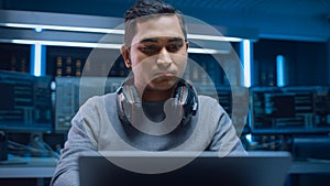 Portrait of Software Developer / Hacker / Programmer Sitting at His Desk and Working on Laptop Com