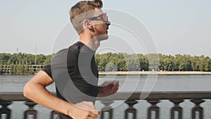 Portrait of sportive athletic boy runs along city river. Side view of slim attractive man in activewear running fast on promenade.