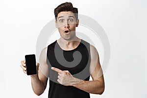 Portrait of sportsman pointing at phone screen with amazed face. Guy in activewear pointing at mobile app interface