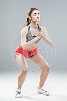 Portrait of sporty athletic woman in sneakers and tracksuit squatting doing sit-ups in gym isolated over gray background