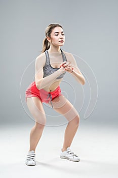 Portrait of sporty athletic woman in sneakers and tracksuit squatting doing sit-ups in gym  over gray background