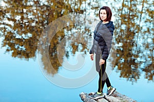 Portrait of sporty beautiful adult brunette woman the fall city park posing near blue lake with autumn trees reflections.