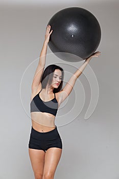 Portrait of sporty woman in black sportswear holding an exercise ball above her head isolated on grey background