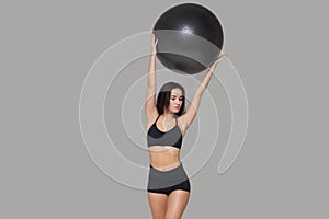 Portrait of sporty woman in black sportswear holding an exercise ball above her head isolated on grey background