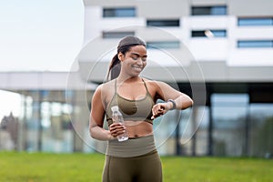 Portrait of sporty young black woman with bottle of water checking her smartwatch or fitness tracker outdoors