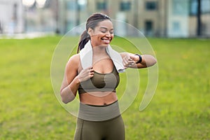 Portrait of sporty young black woman with towel checking her smartwatch or fitness tracker at city park