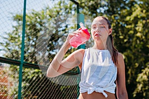 Portrait of sporty young woman drinking cool water from bottle on a summer sports field outdoors