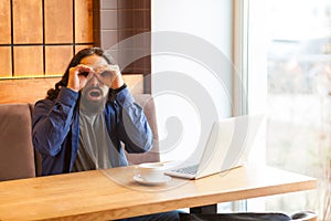 Portrait of spying handsome young adult man freelancer in casual style sitting in cafe with laptop, holding hand near eyes, making