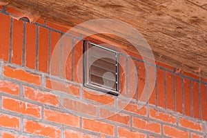 A portrait of a square brown design vent with closed slats placed in a red brick wall on a building for the air output of a cooker