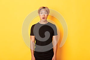 Portrait of startled handsome asian blond guy, drop jaw and gasping impressed, yellow background