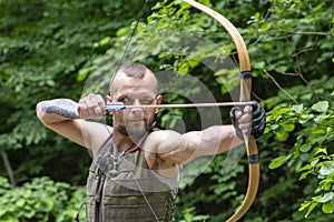 Portrait of a stern man shooting an arrow from a bow in the forest. Concept: men`s hobbies, survival in difficult conditions, hunt