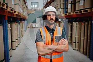 Portrait of storeman in a warehouse for delivering and transporting industrial goods wearing white helmet and orange