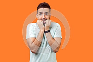 Portrait of stressed out nervous brunette man with beard in white t-shirt biting nails. indoor studio shot isolated on orange