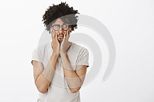 Portrait of stressed and tired handsome male model with tanned skin, rolling eyes in glasses, squeezing cheeks with