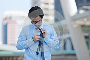 Portrait of stressed young Asian business man binding the tie on urban building in the city background.