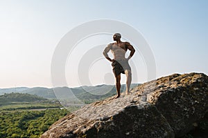 Portrait of strong black african american man bodybuilder with naked torso posing on the rocky peak. Blue cloudy sky