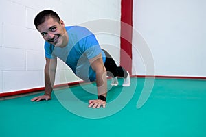 Portrait of a strong determined young man doing push-ups and looking at camera smiling. He is dressed in an orange shirt and a