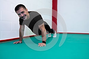 Portrait of a strong determined young man doing push-ups and looking at camera smiling. He is dressed in an orange shirt and a