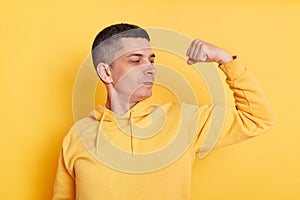 Portrait of strong powerful young adult man wearing casual style hoodie, posing isolated over yellow background, standing with