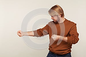 Portrait of strong purposeful man with beard in sweatshirt keeping fists up, holding invisible virtual rope and pulling object