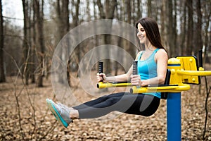 Portrait of strong young woman hanging on wall bars with her legs up. Fitness woman performing hanging leg raises on outdoor in pa