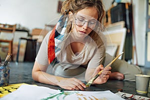 Portrait of a student female artist sitting on the floor in the art studio and painting on paper with a paint brush. A young woman
