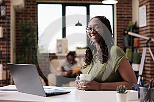 Portrait of student smiling at camera while holding a cup of coffee or tea sitting at desk with laptop