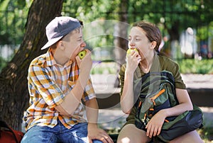 Portrait of students in a city park, teenage schoolchildren a boy and a girl sitting on a bench, resting and eating apples