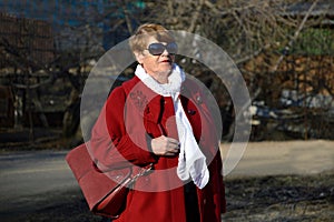 Portrait of stylish 67 year old woman in a bright red coat with a burgundy bag, sunglasses and white scarf on a spring day