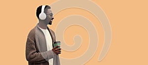 Portrait of stylish african man in wireless headphones listening to music with coffee cup looking away wearing knitted cardigan