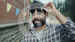 Portrait of stylish Arab guy raising sunglasses smiling and winking outdoors in city street