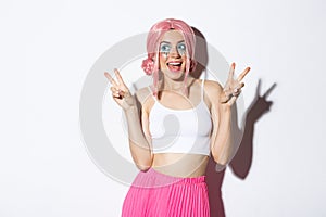 Portrait of stylish attractive girl in pink wig and halloween costume, showing peace signs and smiling happy, standing