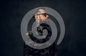 Portrait of a stylish bearded guy with tattooed hands in the military shirt. Studio photo against dark wall