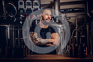 Portrait of a stylish bearded tattooed dark skinned male with crossed arms standing behind the counter in a brewery.