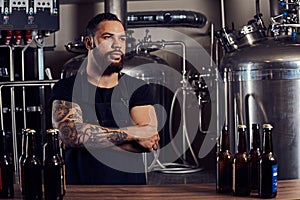 Portrait of a stylish bearded tattooed dark skinned male with crossed arms standing behind the counter in a brewery.