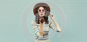 Portrait of stylish beautiful young woman blowing kiss posing in round hat, sunglasses