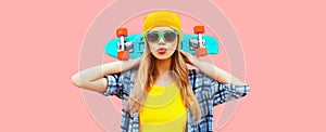 Portrait of stylish blonde young woman model with skateboard wearing colorful yellow hat on pink background