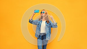 Portrait of stylish blonde young woman taking selfie with smartphone wearing denim jacket on yellow background