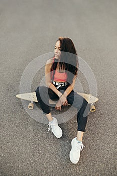 Portrait of stylish brunette girl with curly hair sits on a skateboard on asphalt background, photo from above. Attractive girl