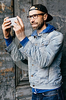 Portrait of stylish guy with beard wearing trendy clothes holding mobile phone making selfie being pleased and smiling with joy in